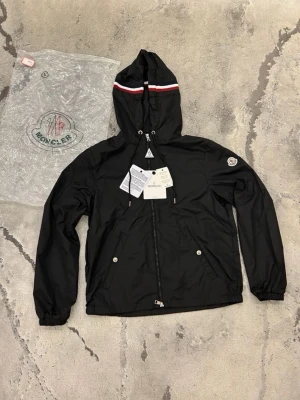 Moncler windbreaker - Snygg svart vindjacka från Moncler med luva och dragkedja framtill. Jackan har vita, blåa och röda detaljer på luvan samt Moncler-logga på armen. Två fickor med knappstängning.