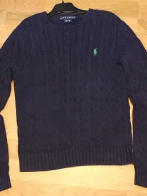 Mörkblå kabelstickad tröja Ralph Lauren - Kabelstickad mörkblå tröja från Ralph Lauren med rund halsringning och grönt broderat logotyp på bröstet. Tröjan har långa ärmar och ribbade muddar vid ärmslut och nederkant. Klassisk och tidlös design i mjukt material.