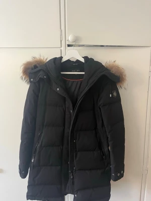 Hollies jacka  - Snygg svart lång dunjacka från Hollies med avtagbar huva och stor fuskpäls i brunt runt huvan. Jackan har dragkedjor framtill, sidofickor med dragkedja och quiltad design. Perfekt för kalla vinterdagar och riktigt varm.