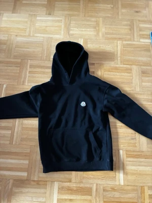 Svart hoodie från Moncler - Snygg svart hoodie från Moncler med klassisk huva och stor magficka. På bröstet sitter Monclers ikoniska logotyp i vitt, rött och blått. Hoodien har ribbade muddar och är gjord i mjukt bomullsmaterial som känns skönt mot huden.