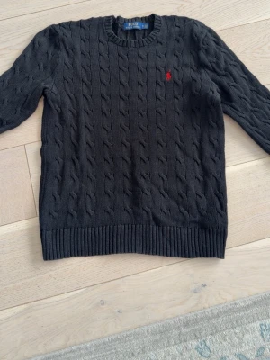 Mörkgrå kabelstickad tröja Polo Ralph Lauren - Kabelstickad mörkgrå tröja från Polo Ralph Lauren med rund halsringning och ribbade muddar. Klassisk röd logga broderad på bröstet. Perfekt till jeans eller kjol för en clean och stilren look.