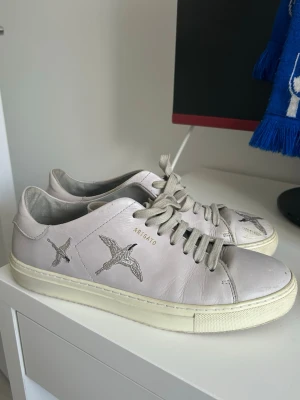 Grå sneakers från Axel Arigato - Säljer ett par grå sneakers från Axel Arigato med silvriga broderade fåglar på sidorna och tån. Skorna har klassisk låg profil, snören och sula i offwhite. Tillverkade i läder för en clean och stilren look. OBS: lädret har skadats på tårna, se bild 2.
