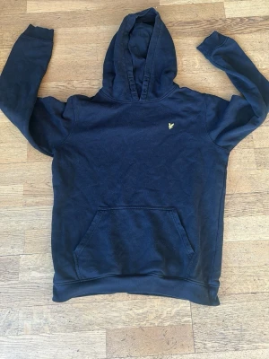 Svart hoodie från Lyle & Scott - Klassisk svart hoodie från Lyle & Scott Junior med gul broderad logga på bröstet och stor känguruficka framtill. Hoodien har en mjuk insida och ribbade muddar vid ärmar och nederkant. Perfekt för en avslappnad och stilren look.