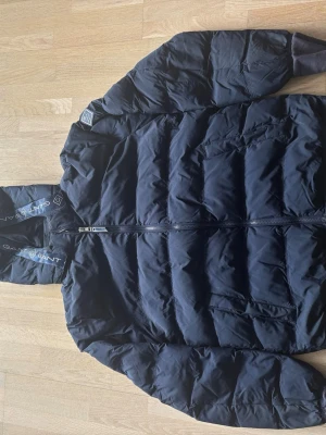 Svart dunjacka från Gant med huva - Snygg svart dunjacka från Gant med huva och dragkedja framtill. Jackan har broderad logga på huvan och är quiltad med breda kanaler. Perfekt för kalla vinterdagar och har en modern, sportig look. Jag har lagt till en liten extra del på armarna så att den blir lite längre vid armarna.
