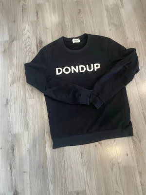 Svart sweatshirt från Dondup - Säljer en svart sweatshirt från Dondup med vit logga tryckt över bröstet. Tröjan har rund halsringning, långa ärmar och ribbade muddar vid ärmslut och nederkant. Perfekt för en clean och stilren look.
