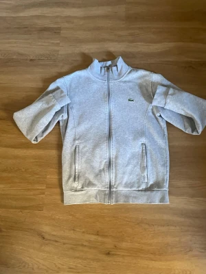 Grå zip hoodie från Lacoste - Snygg grå zip hoodie från Lacoste med klassisk krokodil-logga på bröstet. Tröjan har hög krage, dragkedja framtill och två fickor. Tillverkad i mjukt sweatshirtmaterial som är riktigt skönt att ha på sig. Perfekt för en chill och sportig look.