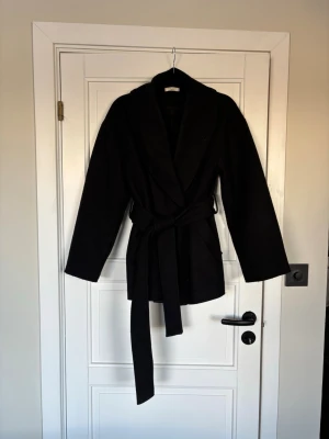 Svart kappa med knytskärp - Stilren svart kappa från Ginatricot med bred krage och knytskärp i midjan. Kappan har fickor och knappar. Modellen är midjelång och har raka ärmar. Perfekt för dig som gillar en enkel men trendig look. 