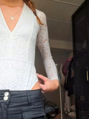 Vit spetsbody med lång ärm - Snygg vit body i spets med långärmad design och djup V-ringning. Kroppsnära passform och transparenta ärmar ger en trendig look. Perfekt att styla med jeans eller kjol för en chic outfit.