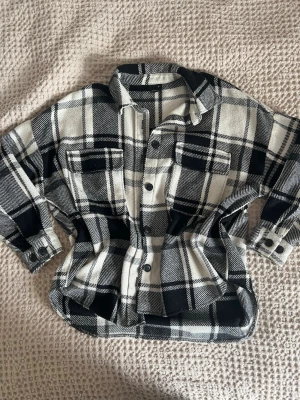 Svartvit rutig mjuk jacka - Svartvitt rutigt mönster med knappar framtill och på ärmsluten. Två bröstfickor med lock och en mjuk, lite tjockare vävd kvalitet som känns perfekt för lager på lager. Klassisk krage och avslappnad passform.