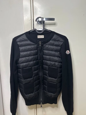 Svart cardiganjacka från Moncler - Snygg svart cardiganjacka från Moncler med quiltad front och stickade ärmar. Två framfickor i ribbstickat material och dragkedja framtill. Rund halsringning och Moncler-logga på ärmen. Perfekt för lager på lager och streetstyle.