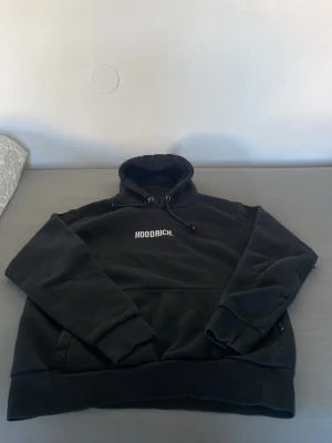 Svart hoodie från Hoodrich - Säljer en svart hoodie från Hoodrich med vit logotyp på bröstet. Hoodien har en stor huva med dragsko, ribbade muddar och en praktisk magficka. Perfekt för dig som gillar streetwear och vill ha något bekvämt och stilrent. Inga fläckar på tröjan 