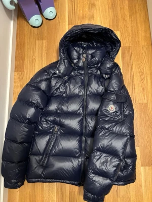 Mörkblå Moncler pufferjacka med huva - Säljer en mörkblå pufferjacka från Moncler med huva och dragkedja framtill. Jackan har Moncler-logga på ärmen, två snedställda fickor med dragkedja och blanka knappar. Perfekt för kalla dagar och riktigt snygg streetstil.