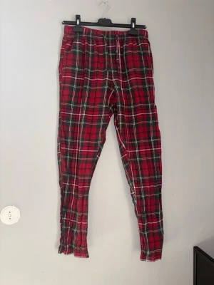 Rödrutiga pyjamasbyxor - Säljer ett par klassiska pyjamasbyxor i röd, grön och vit rutigt mönster. Byxorna har resår i midjan och är i mjukt, tunt tyg som passar perfekt för att chilla hemma. Perfekta för mysiga kvällar eller lata morgnar.