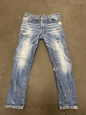 Blå Dsquared 2 jeans med färgstänk - Säljer ett par blå jeans med slitningar, hål och coola färgstänk i flera färger. Jeansen har skinny fit, klassiska fem fickor och dragkedjegylf. Materialet är denim i bomull och de har en trendig, använd look. Mellan M och S. Passar båda två. 