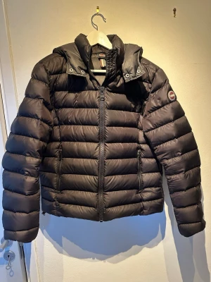Svart dunjacka från Colmar - Snygg svart dunjacka från Colmar med huva och dragkedja framtill. Jackan har två sidofickor med dragkedja och klassisk quiltad design. Perfekt för vår vädret. Den är inte så tjock som den ser ut och är riktigt skön att bära. Märkeslogga på ärmen och diskret logga bak.