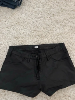 Skinnshorts - Snygga svarta shorts från Bik Bok i skinnimitation. Klassisk femficksmodell med bälteshällor och knapp- samt dragkedjestängning. Perfekta för dig som vill ha en edgy vibe till din outfit.