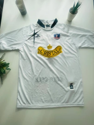 Vit Colo-Colo fotbollströja 💫🔥 - Säljer en vit fotbollströja från Colo-Colo med svart krage och broderad klubbmärke på bröstet. Tröjan har stor gul Cristal-logga på magen och PENKO-logga på axeln. Perfekt för dig som älskar fotboll och vill representera ditt lag.