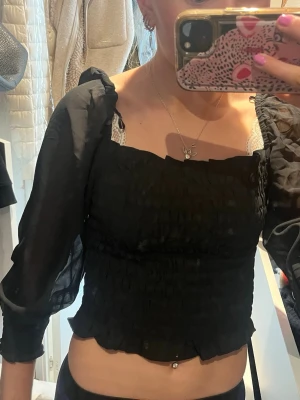 Svart croppad topp med puffärm - Trendig svart croppad topp med transparenta puffärmar och smockad kropp. Fyrkantig urringning och lätt volangkant nedtill ger en snygg siluett. Perfekt för dig som vill ha en statement topp till din outfit.