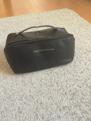 Svart necessär Rose of Na Mon's Land - Snygg svart necessär med guldfärgad text på framsidan. Väskan har ett praktiskt handtag upptill och dragkedja runt om. Perfekt för att organisera smink eller småprylar. Tillverkad i ett slätt, tåligt material.