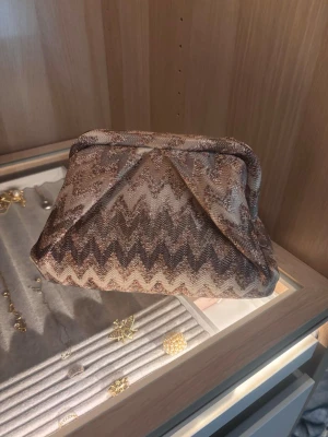 Becksöndergaard väska beige  - Säljer en snygg Becksöndergaard väska i beige och bruna toner med ett coolt zigzag-mönster. Väskan har en mjuk textilstruktur och stängs upptill. Insidan är fodrad i brunt tyg och har en liten innerficka med dragkedja. 