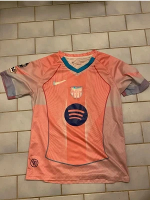 Barcelona matchtröja rosa Nike - Säljer en rosa Barcelona matchtröja från Nike med klubbmärke och sponsorlogga på bröstet. Tröjan har turkos V-ringad krage, vita ränder och detaljer i lila. Perfekt för dig som älskar fotboll och vill sticka ut på planen.