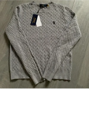 Grå kabelstickad tröja Polo Ralph Lauren - Snygg grå kabelstickad tröja från Polo Ralph Lauren med rund halsringning och diskret broderad logga på bröstet. Tröjan har ribbade muddar vid ärmslut och nederkant. Perfekt för klassisk stil.