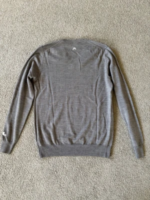 Grå V-ringad tröja från J.Lindeberg - Snygg grå V-ringad tröja från J.Lindeberg med diskret logga på ryggen och ärmen. Tröjan har långa ärmar och ribbade muddar. Perfekt för lager-på-lager och passar till många olika outfits.