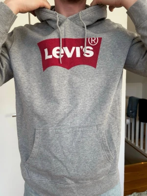 Grå Levi's hoodie med logga - Säljer en grå hoodie från Levi's med stor röd och vit logga på bröstet. Hoodien har huva med dragsko och en stor magficka framtill. Tillverkad i mjukt sweatshirtmaterial