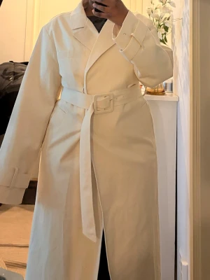 Beige trenchcoat från NA-KD - Stilren beige trenchcoat från NA-KD med klassisk krage, knytskärp i midjan och dubbla knappar framtill. Långa ärmar med spänndetaljer vid handlederna och två stora fickor. Perfekt för en trendig och avslappnad look.