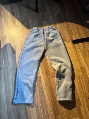 Grå mjukisbyxor med logga - Säljer ett par grå mjukisbyxor med resår i midjan och sidofickor. Byxorna har en vit broderad logga på vänster sida och dragkedjor vid bensluten. Perfekta för chill dagar eller när du vill vara bekväm.