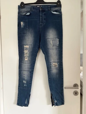 Blå slitna jeans med dragkedjor killbyxor  - Herrbyxor - Säljer ett par blå jeans med slitningar och patchade detaljer på benen. Jeansen har dragkedjor vid bensluten och en tvättad look som ger en cool och avslappnad stil. Perfekta för dig som gillar streetwear och vill ha något unikt i garderoben. Använda 1-2 gånger 