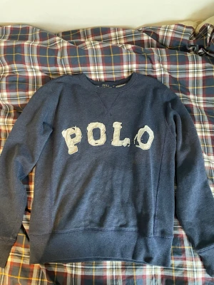 Blå sweatshirt från Polo Ralph Lauren - Mörkblå sweatshirt från Polo Ralph Lauren med stor vit POLO-text framtill. En perfekt tröja för mer skön stil. 
