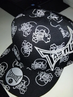 Svart Tapout keps med dödskallemönster - Säljer en svart keps från Tapout med broderad logga framtill och vitt dödskallemönster över hela kepsen. Klassisk flatbrätte och cool streetwear-stil. Märket Tapout syns tydligt både på framsidan och på klistermärket på skärmen.