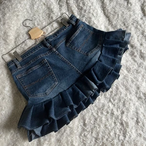 Kort jeanskjol med volanger - Kort jeanskjol med dubbla volanger! Längd bak: 34cm, fram: 26cm och midjemåttet vid knapphöjden är 39cm. 