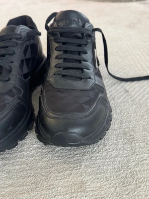 Svarta sneakers från Prada - Snygga svarta sneakers från Prada med diskret mönstrad textil och läderdetaljer. Klassisk låg modell med svarta skosnören och ikonisk metallogo på sidan. Perfekta för dig som gillar stilrena och exklusiva sneakers. Med kvitto  (köpte nypris 10600kr) 
