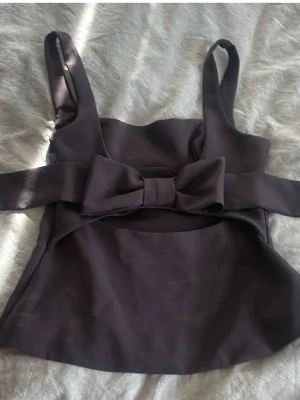 Topp Zara  - Snygg svart linnetopp med breda axelband och en stor rosett framtill. Toppen har en figurnära passform och är tillverkad i ett mjukt, något stretchigt material. Perfekt att styla med jeans eller kjol för en trendig look.