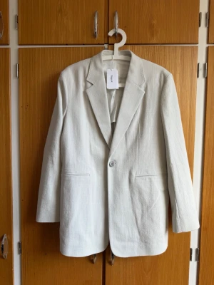 Vit kavaj från Filippa K - Stilren vit kavaj från Filippa K med klassisk passform, enkelknäppt framtill och två fickor. Tillverkad i ett strukturerat tyg med slät fodrad insida. Perfekt för en clean och modern look.