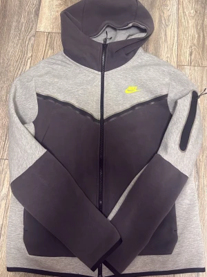 Nike Tech Fleece hoodie grå/svart - Nike Tech Fleece hoodie i grått och svart med hel dragkedja och huva. Snygg kontrastdesign, neon grön Nike-logga på bröstet och praktisk ficka med dragkedja på ärmen. Perfekt för en sportig och modern look. Pris kan diskuteras.