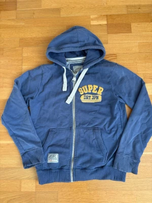 Blå zip hoodie från Superdry - Blå hoodie med dragkedja från Superdry. Har huva med vita snören, ribbade muddar och fickor framtill. Broderad gul och vit Superdry-logga på bröstet samt patch vid fickan. Perfekt för en avslappnad och cool stil.