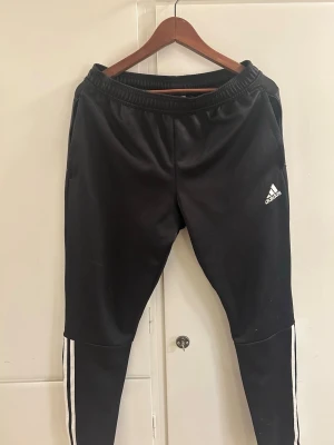Svarta Adidas träningsbyxor M - Säljer ett par svarta träningsbyxor från Adidas med klassiska vita ränder längs benen och Adidas-logga på vänster lår. Byxorna har resår i midjan och är tillverkade i ett lätt och stretchigt material som passar perfekt för träning eller chill.