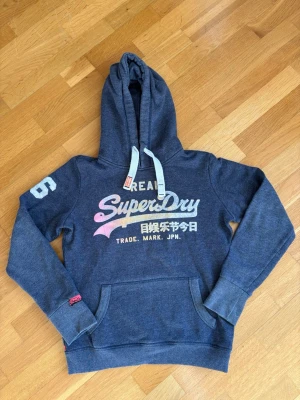 Blå Superdry hoodie med tryck - Mörkblå hoodie från Superdry med stor logga och japansk text på bröstet. Har vit dragsko i huvan, magficka och tryckt siffra på ärmen. Mjuk bomullsblandning och ribbade muddar. Perfekt för en avslappnad streetstyle.