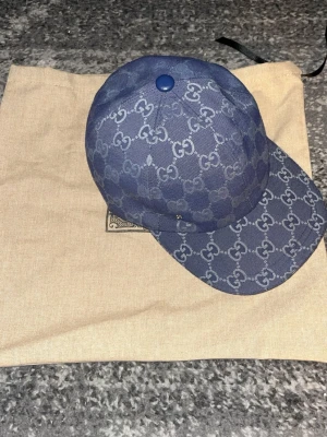 Blå keps från Gucci med GG-mönster - Säljer en snygg blå keps från Gucci med det klassiska GG-monogrammet över hela kepsen. Kepsen har böjd skärm och är tillverkad i ett slitstarkt tygmaterial. Perfekt för dig som vill ha en exklusiv och trendig accessoar.