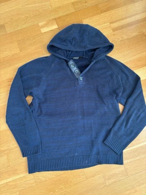 Mörkblå hoodie från livergy  - Mörkblå stickad hoodie från livergy med huva och tre knappar vid halsen. Tröjan har långa ärmar, ribbade muddar och nederkant. Perfekt för dig som gillar en enkel men snygg stil. Materialet känns mjukt och bekvämt.