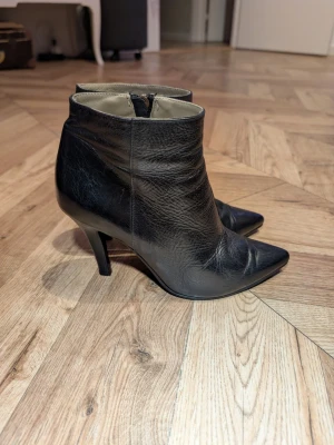 Svarta spetsiga stövletter i skinn - Snygga svarta stövletter i äkta skinn med spetsig tå och smal klack. Dragkedja på insidan för enkel påtagning. Stilren design som passar till många outfits. Perfekta för dig som gillar en klassisk och trendig look.