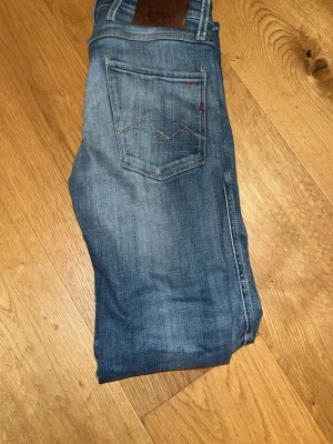 Blå slitna jeans från Replay KAN BYTA - Säljer ett par blå jeans från Replay med snygga slitningar och detaljer på både fram- och bakfickor. Jeansen har klassisk femficksdesign, normal midja och är tillverkade i denim med lätt stretch. Perfekta för en avslappnad och trendig look.