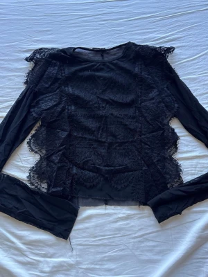 Svart spetsblus med lång ärm - Snygg svart blus med lång ärm och transparent spetsdetaljer längs sidorna och axlarna. Blusen har volangliknande spets 
