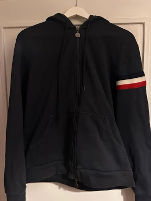 Blå Moncler hoodie med dragkedja - Blå hoodie från Moncler.. På ena ärmen finns en stickad röd och vit rand samt Moncler-logga. Perfekt för en chill och stilren look.