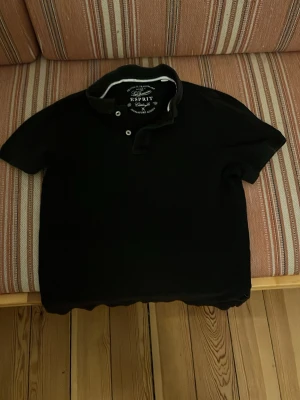 Svart pikétröja från Esprit - Klassisk svart pikétröja från Esprit med krage och knappar framtill. Tröjan har korta ärmar och är tillverkad i ett mjukt bomullsmaterial som känns skönt mot huden. Perfekt till jeans eller shorts för en enkel och stilren look.