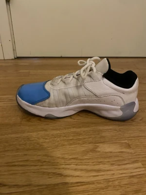 Jordan sneakers vit/blå - Snygga Jordan sneakers i vitt och blått med svarta detaljer. Skorna har lågt skaft, vit ovandel i läder och mesh, samt blå lackad tå och sidor. Klassisk Jumpman-logga på hälen. Perfekta för dig som gillar streetwear och sportig stil. Använda få gånger. Kan användas utomhus och inomhus.