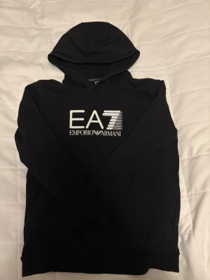 Emporio Armani Hoodie  - Säljer en svart och varm Emporio Armani hoodie med stort tryck på bröstet och en ficka med dragkedja. Mer information är bara att fråga om.